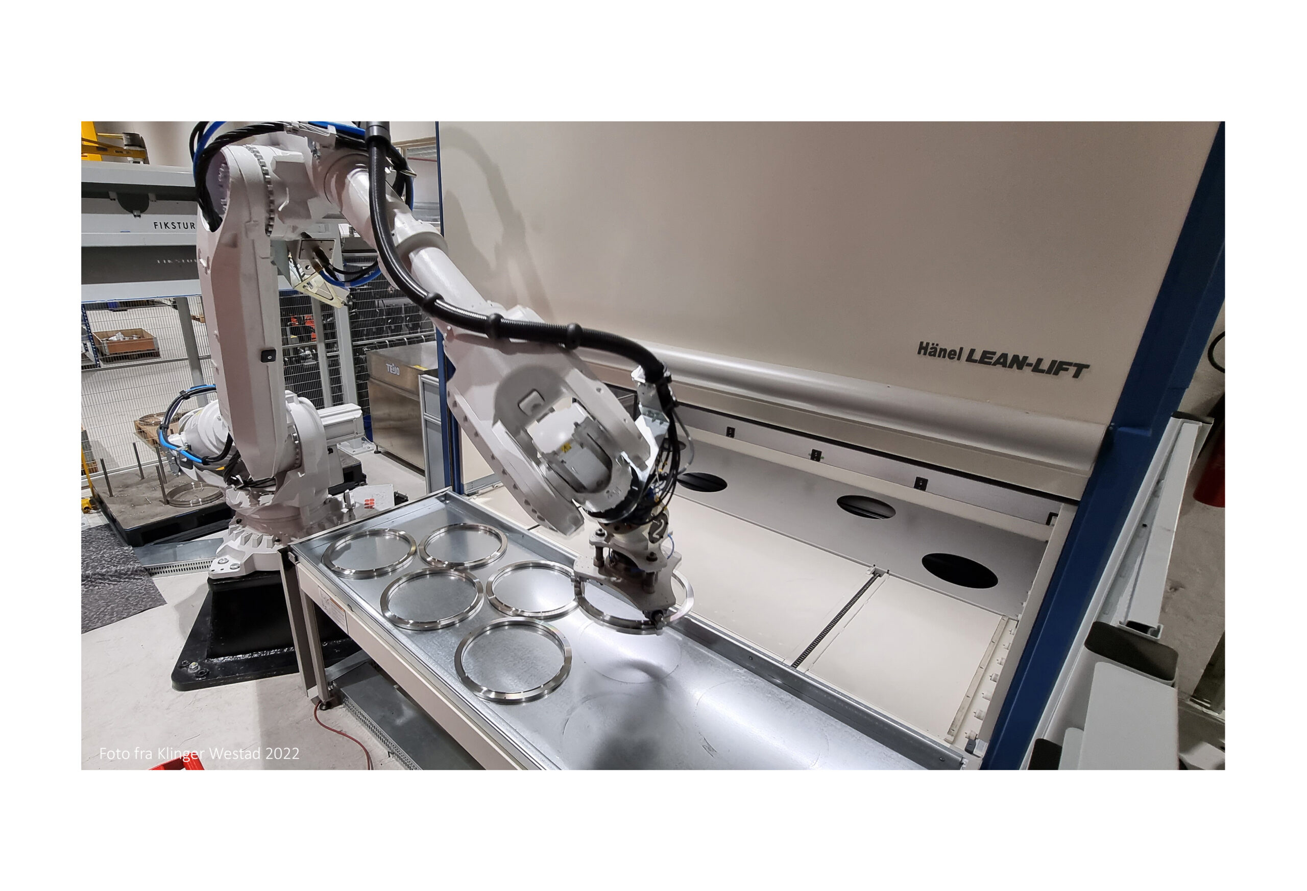 Industrial robotic arm handling metal discs on a tray in a Lean-Lift manufacturing cell, with a large white unit labeled Hänel LEAN-LIFT in the background.