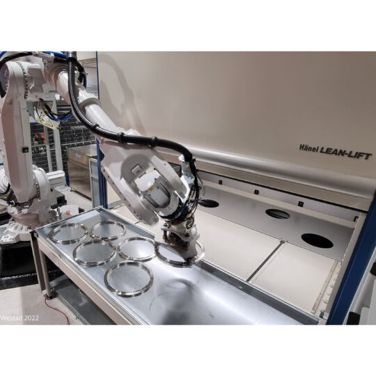 Industrial robotic arm handling metal discs on a tray in a Lean-Lift manufacturing cell, with a large white unit labeled Hänel LEAN-LIFT in the background.