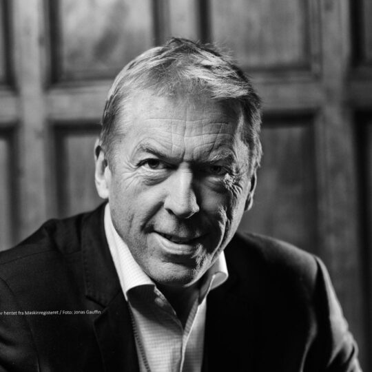 Black-and-white close-up portrait of a smiling middle-aged man in a suit, looking at the camera with a wooden background behind him.
