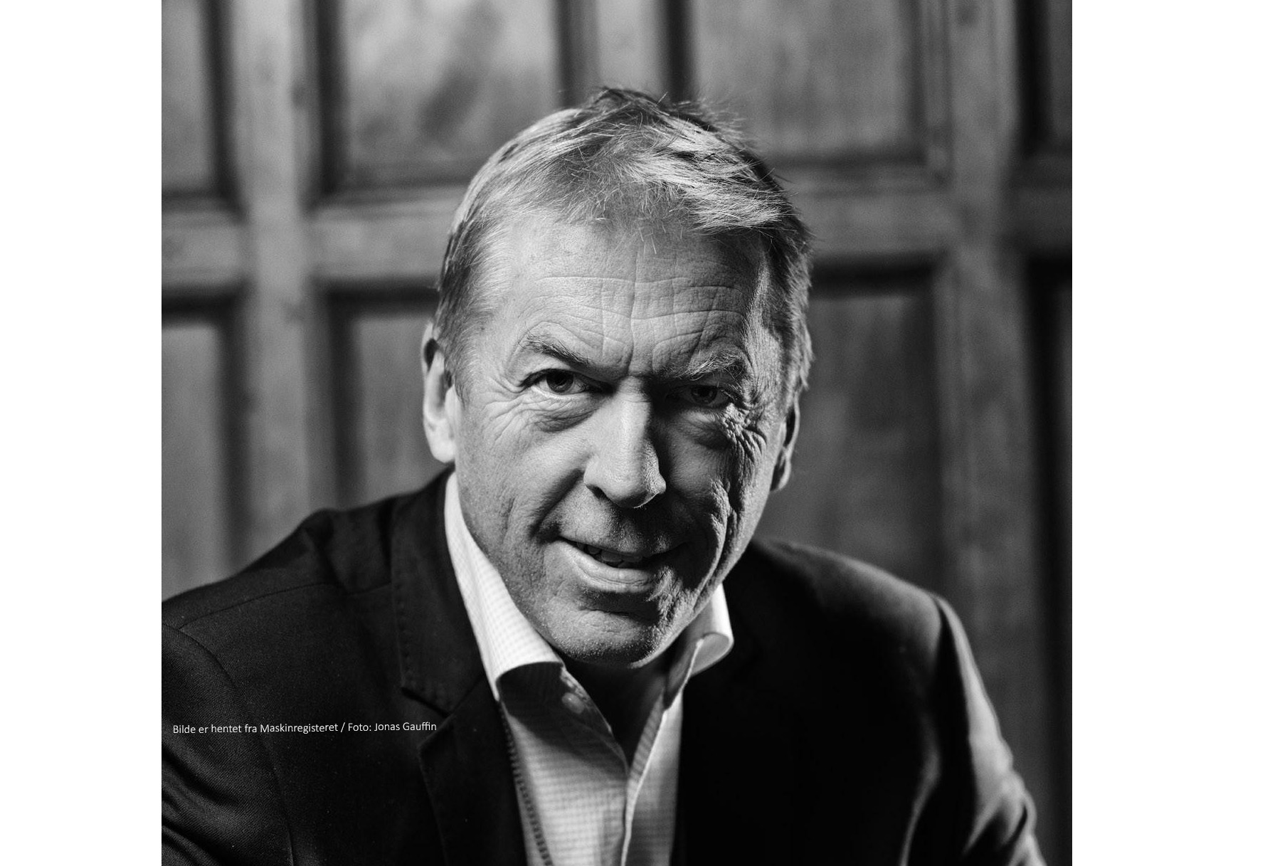 Black-and-white close-up portrait of a smiling middle-aged man in a suit, looking at the camera with a wooden background behind him.