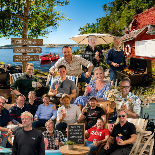 Group of people at a seaside barbecue, smiling around drinks and a grill, with wooden signs and a red building nearby.